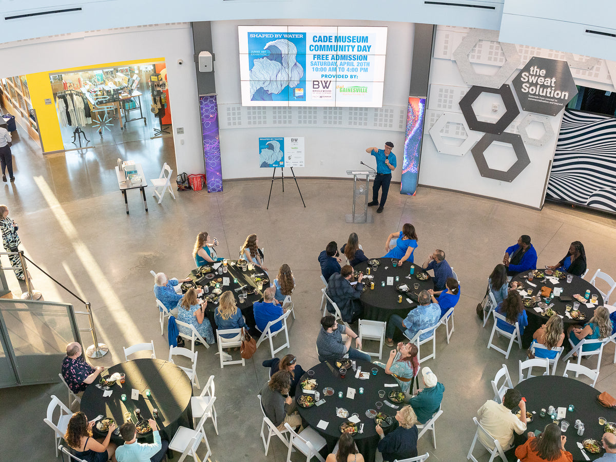 A high-angle view of a community event at the Cade Museum, showing a presenter at a podium and a group of people seated at round tables with food and drinks. A large screen displays 'Cade Museum Community Day'.