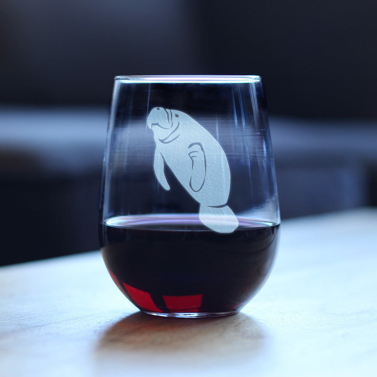 Stemless wine glass with a manatee etching on it, filled with red wine and sitting on a wooden table.