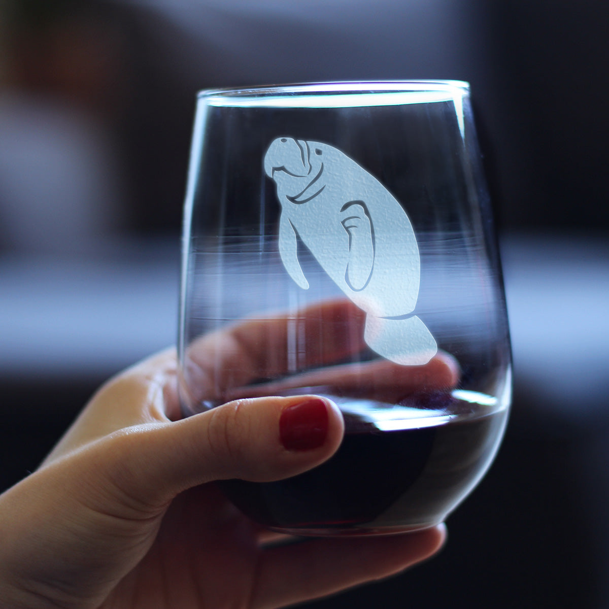 A hand holding a stemless wine glass with an etched manatee design on it against a blurred background.