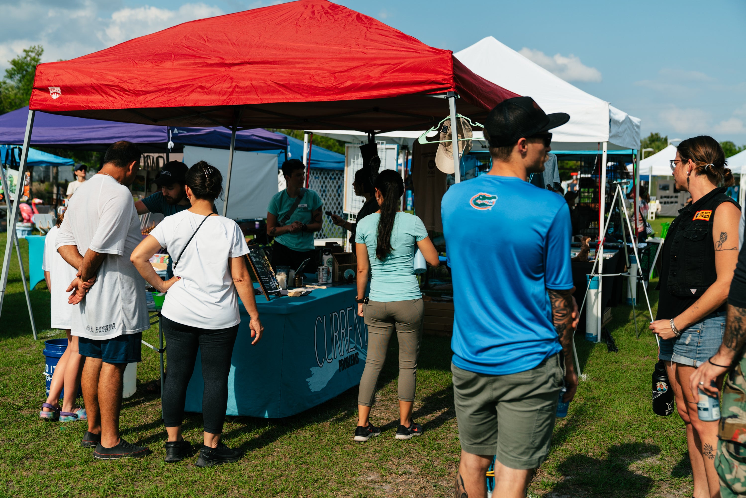 A group of people standing and browsing at an outdoor market booth under a red canopy tent on a grassy field.