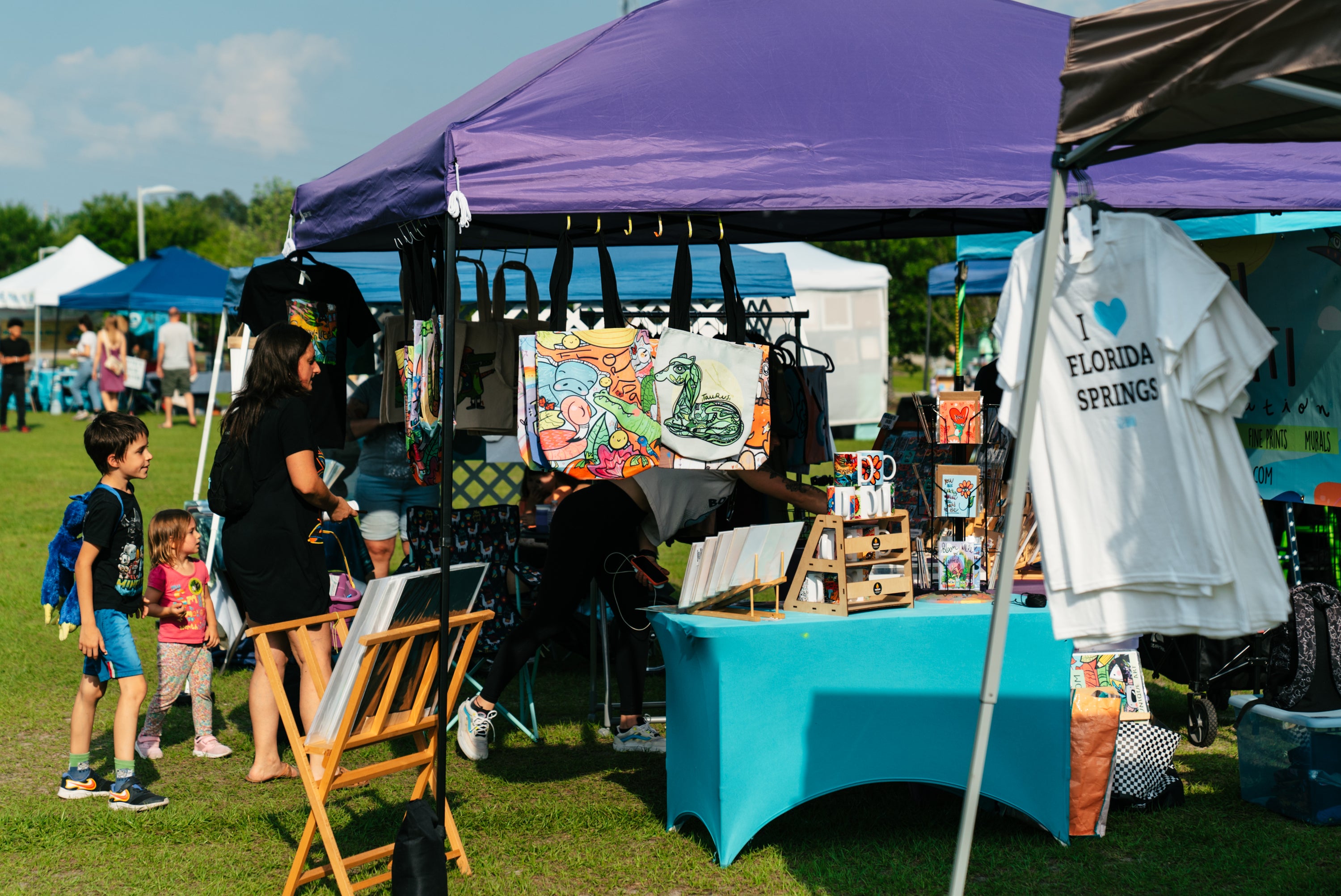 An outdoor market scene under purple and black canopy tents, with a table displaying handmade items, a white T-shirt hanging on the right that reads 'I LOVE FLORIDA SPRINGS', and several people standing nearby on a grassy field.