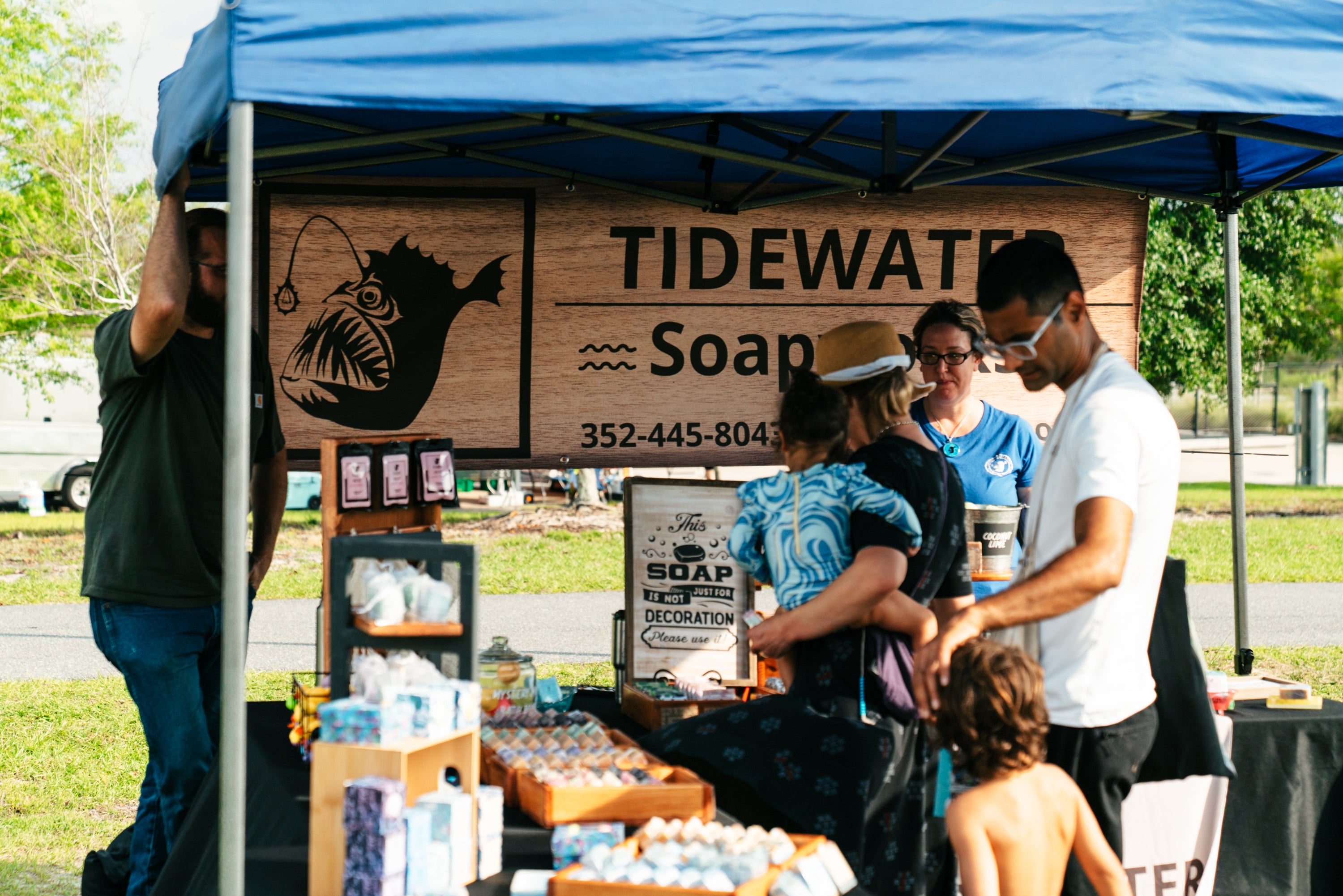 A pop-up canopy booth at an outdoor market with a large light-brown banner reading 'TIDEWATER Soap' and an anglerfish graphic. Several people are gathered around the booth, and soap products are displayed on the table below.