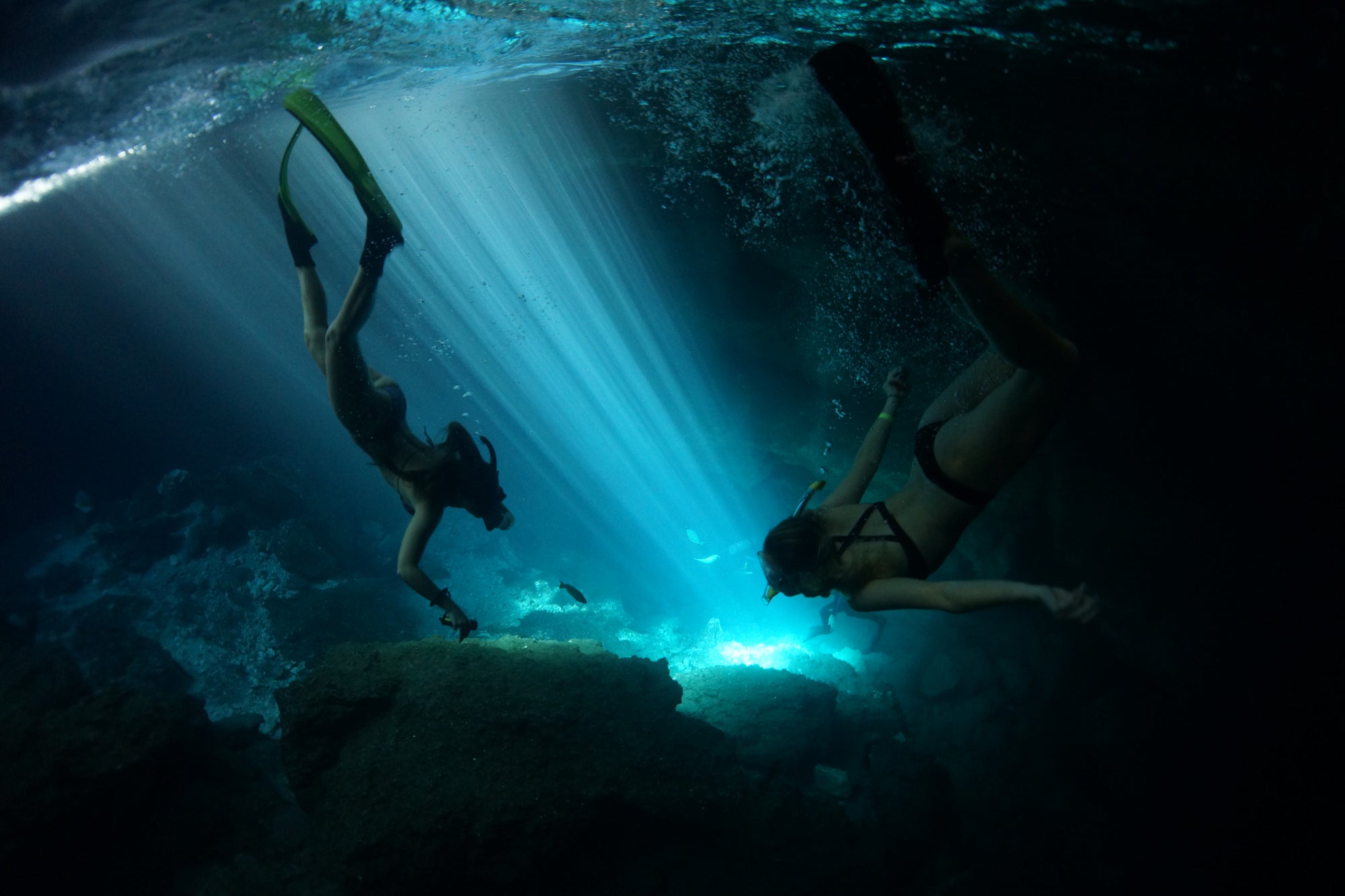 Two women snorkeling underwater in a dark blue cave, with bright sunbeams filtering through the surface and turquoise light reflecting off rocks below.
