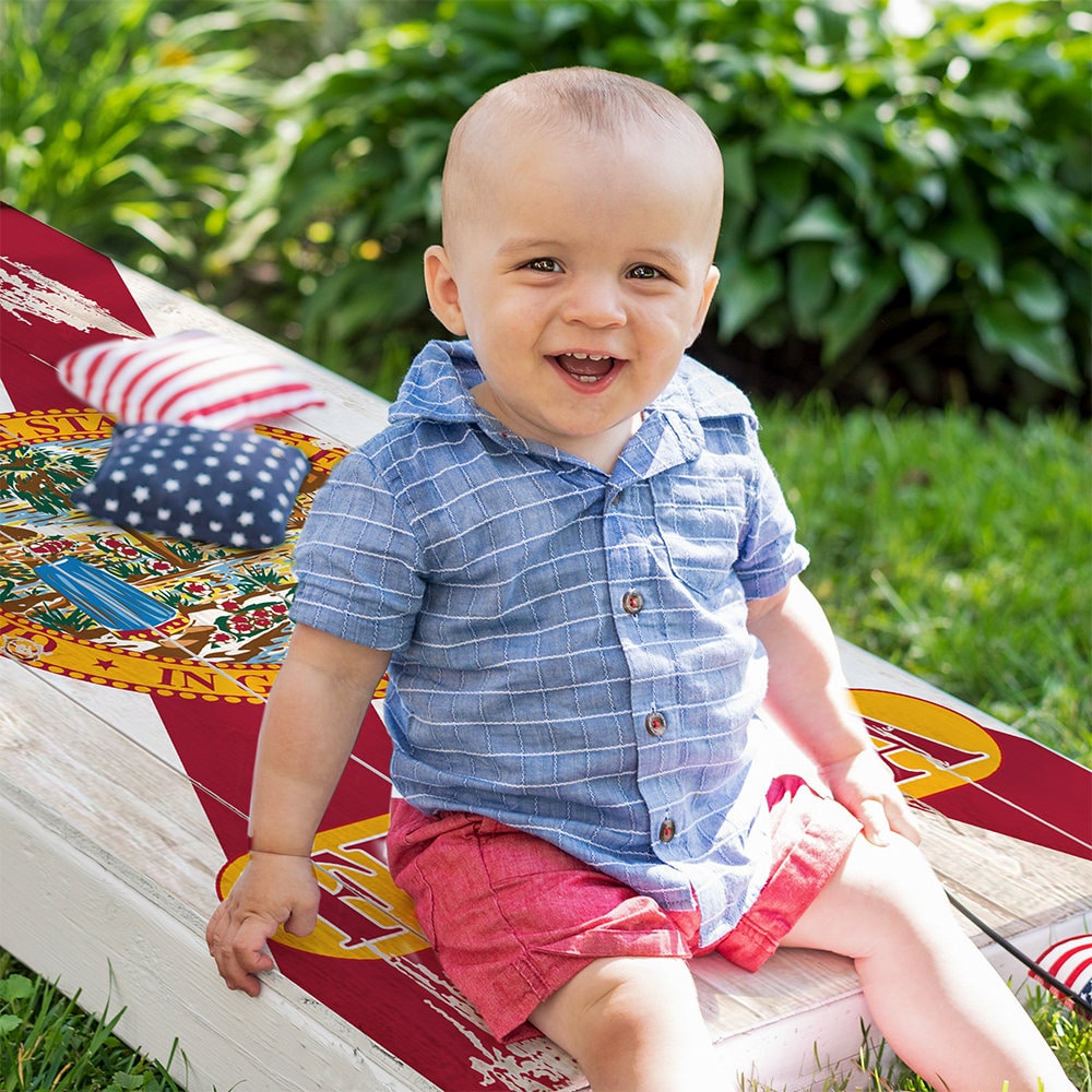 A baby in a blue shirt and red shorts holding a small American flag sitting on a cornhole board with a Florida flag design on it. There are also American flag patterned cornhole bags on the board.