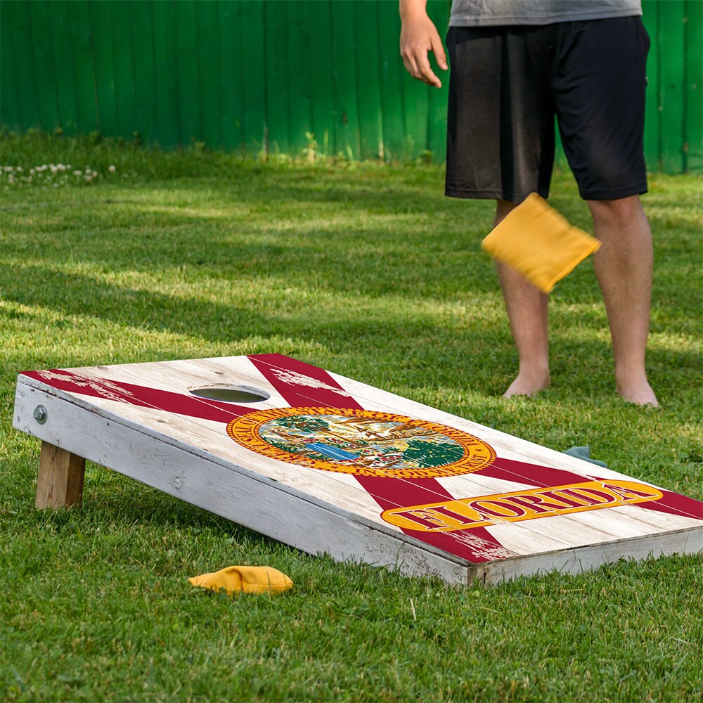 Person playing cornhole on a grassy area with a green fence in the background. They are using a cornhole board with a Florida map design on it.