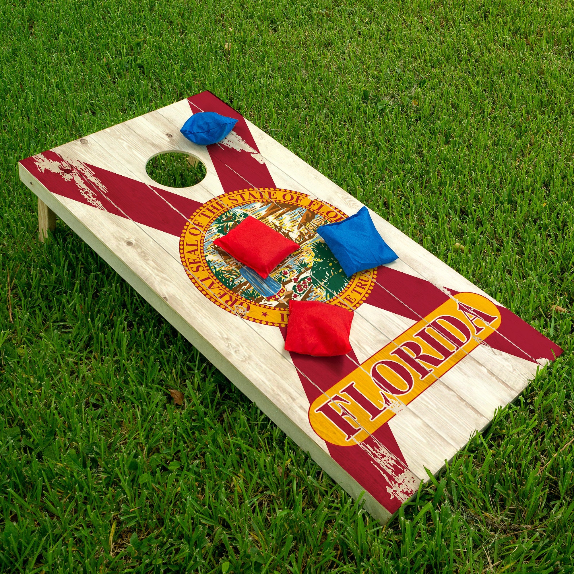 A cornhole board with a Florida flag design on a grassy field, with red and blue cornhole bags on top of it.