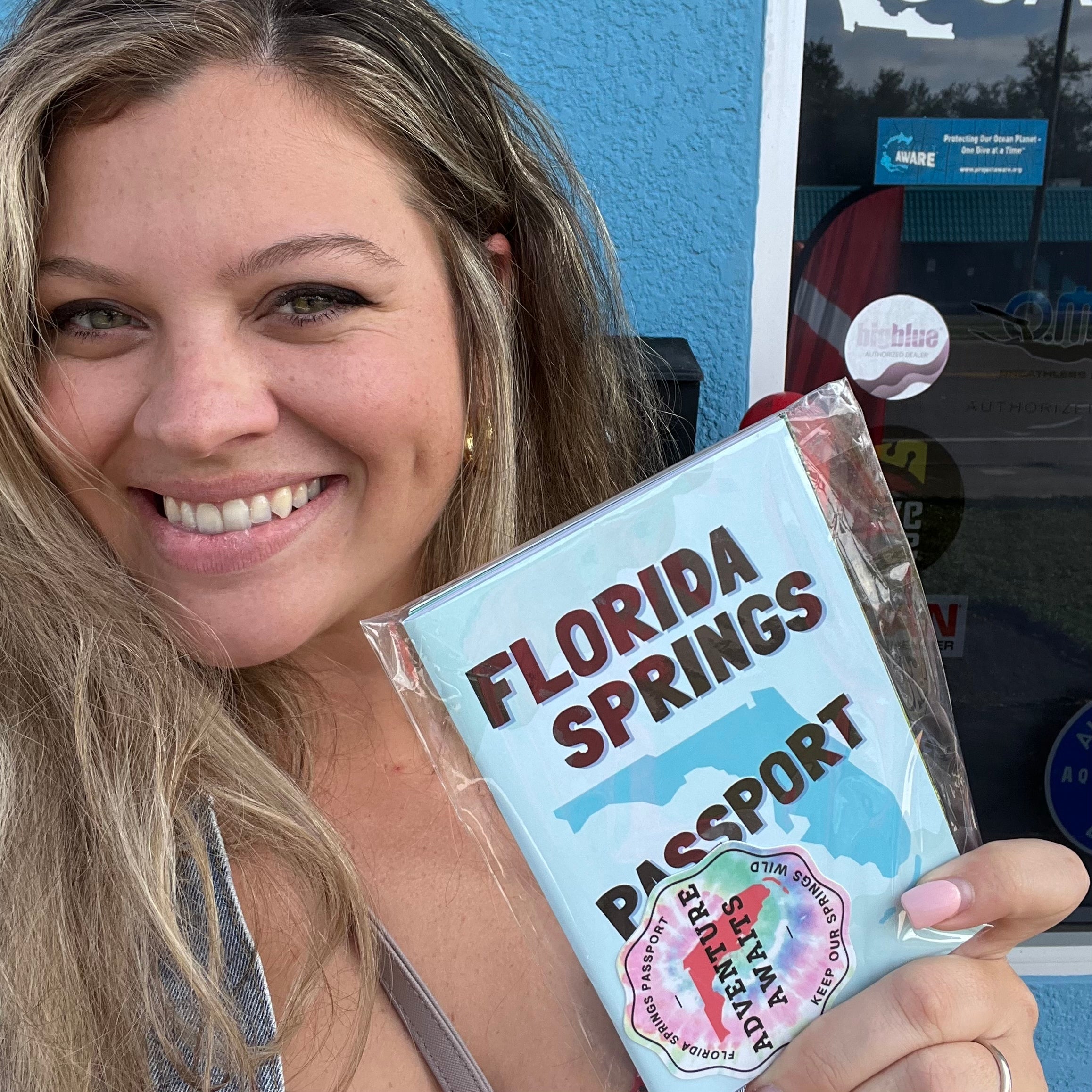 A smiling woman (Danielle) with long light brown hair holds a clear plastic-wrapped novelty passport labeled 'Florida Springs Passport' in front of a blue building wall.