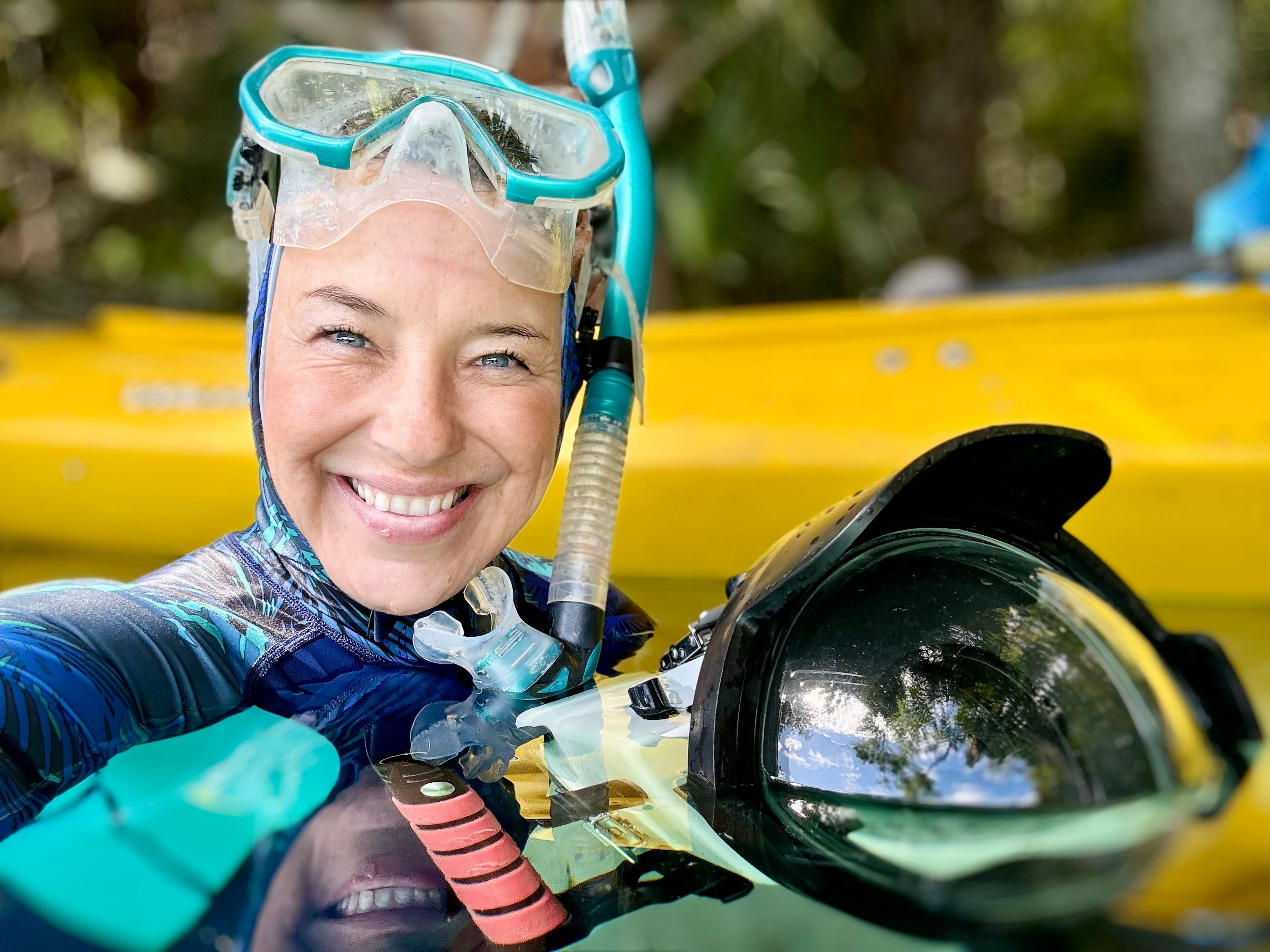 A smiling woman (Linda) wearing a blue wetsuit, turquoise snorkeling mask, snorkel, and holding a large underwater camera in front of a yellow boat with a blurred outdoor background.